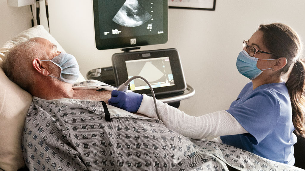 Paciente sometiéndose a un ecocardiograma mientras observa el monitor de ultrasonido y un médico sonriente.